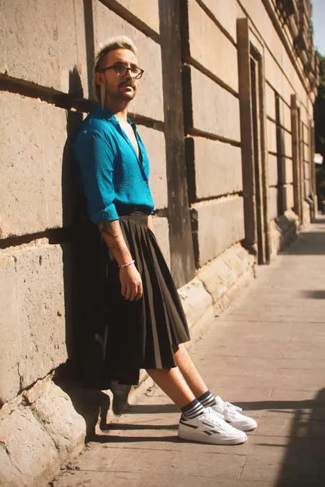 Hombre con gafas, camisa turquesa y falda negra plisada apoyado en un muro, con zapatillas blancas