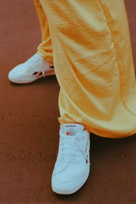 Detalle de pantalón amarillo holgado y zapatillas blancas Reebok sobre fondo rojizo