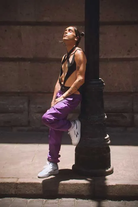 Persona con pantalón morado y zapatillas blancas apoyada en un poste, mirando hacia arriba en una calle