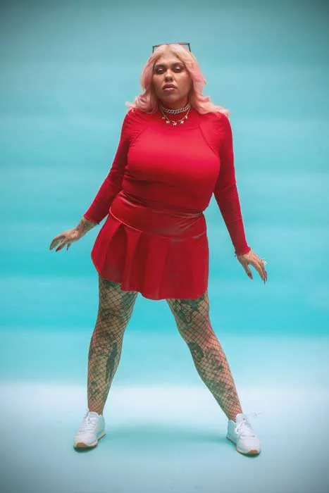 Persona de cabello rosado con vestido rojo, piernas tatuadas y zapatillas blancas, sobre fondo turquesa