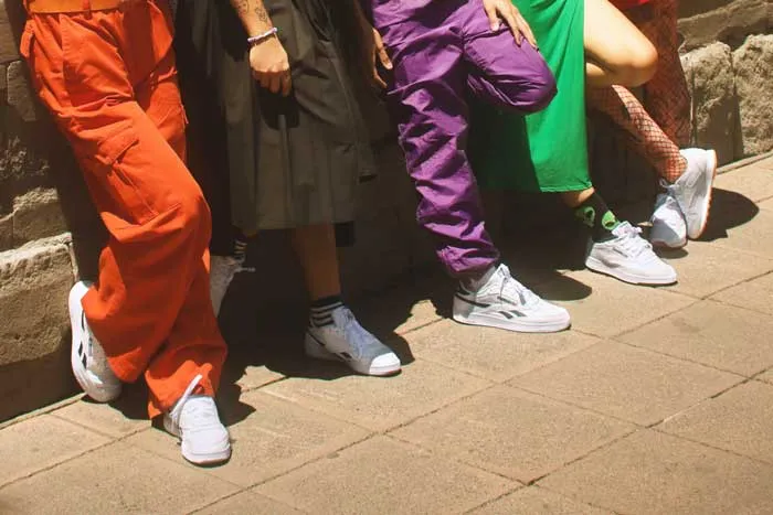 Piernas de varias personas con pantalones naranja, morado y verde, falda negra y zapatillas blancas, sentadas en la acera
