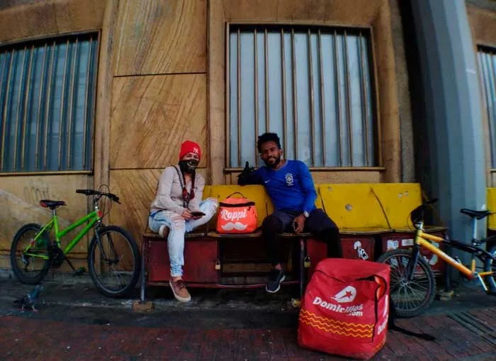Dos repartidores con mascarilla descansan sentados en una banca junto a bicicletas y una mochila roja de domicilios