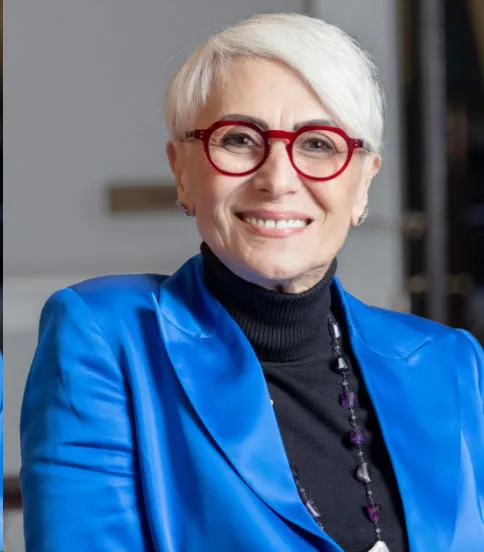 Retrato de una mujer de cabello blanco con gafas rojas, saco azul y cuello alto negro, sonriendo
