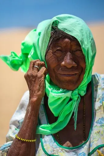 Retrato de una mujer mayor con pañuelo verde brillante, que se toca el rostro y mira a la cámara