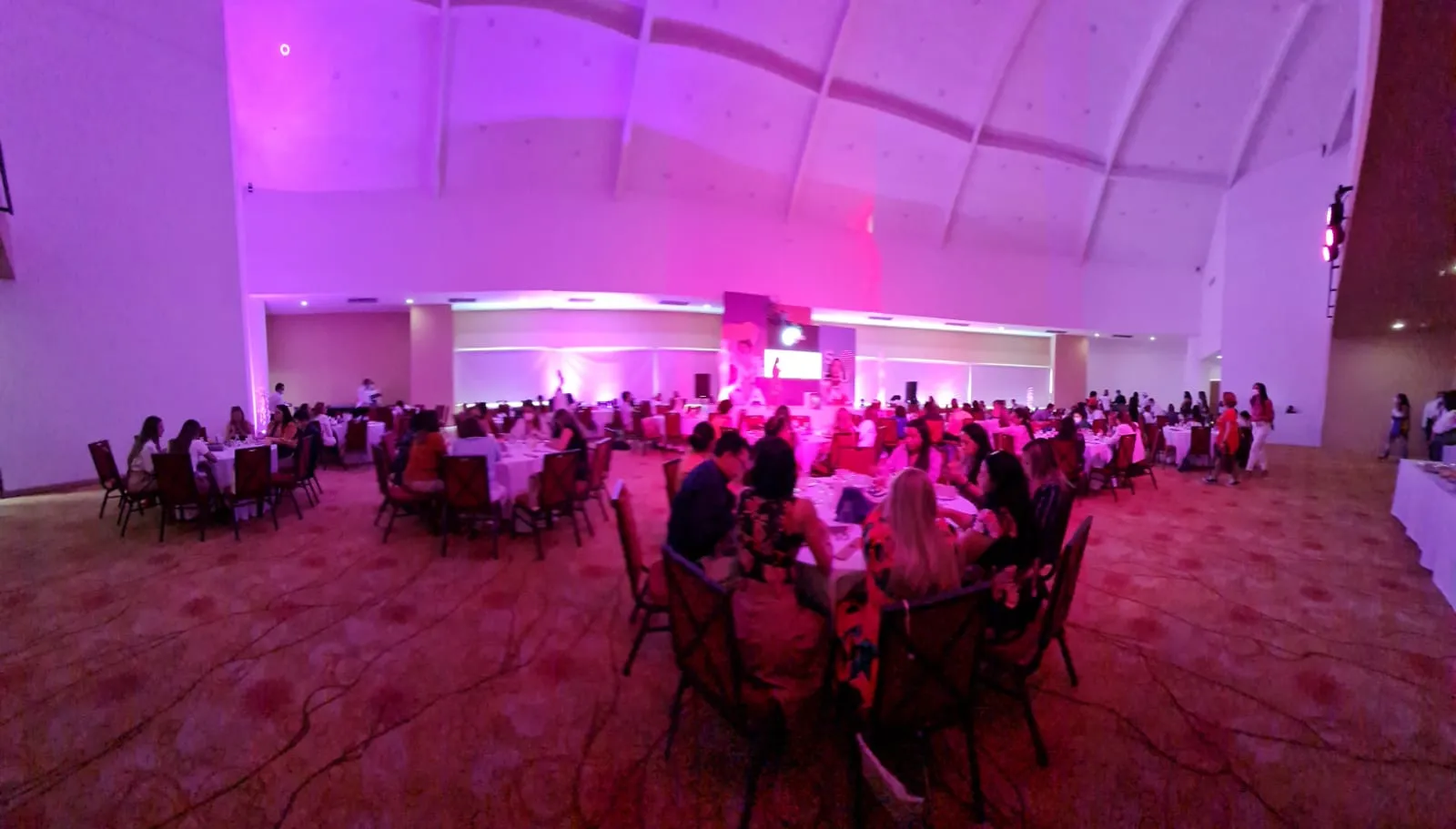 Salón de banquetes iluminado en tonos rosa y morado con asistentes sentadas a las mesas