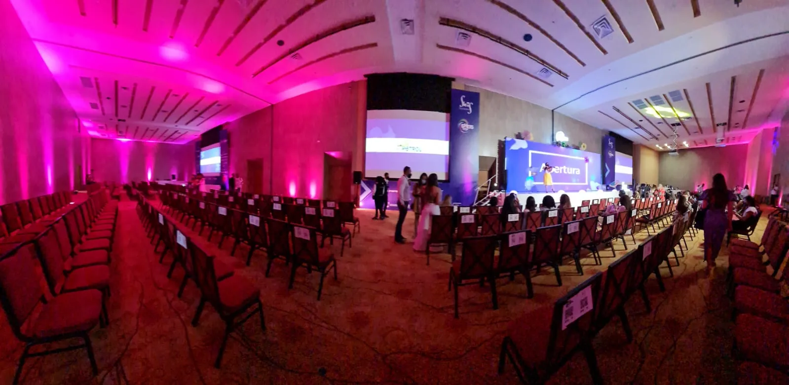 Panorámica de un salón de eventos iluminado en rosa con filas de sillas y mesas para el She is Global Forum