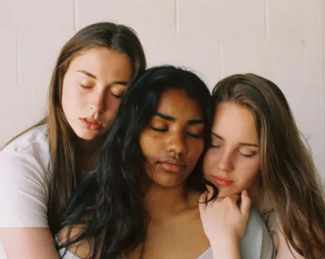 Tres mujeres jóvenes con los ojos cerrados apoyan sus cabezas unas contra otras
