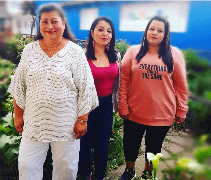 Tres mujeres posan de pie en un jardín, una mayor con blusa blanca y dos con buzos de colores