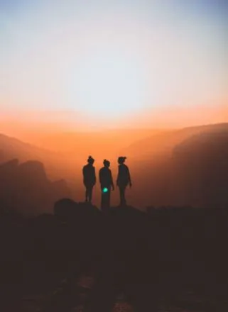 Silueta de tres personas de pie sobre una cima frente a un atardecer anaranjado entre montañas