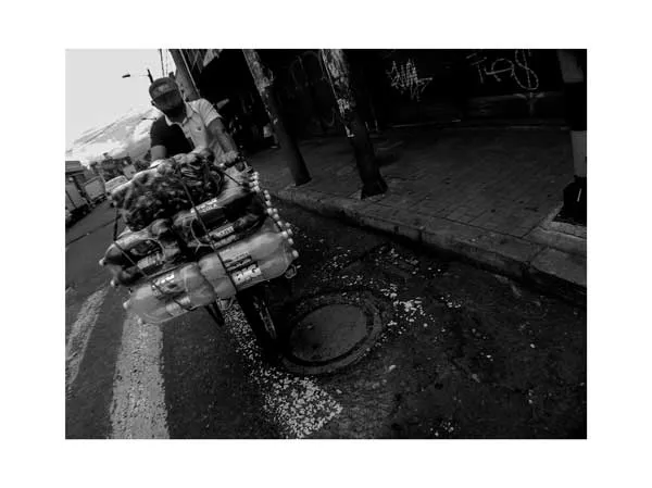 Vendedor ambulante empuja un carrito cargado de mercancía por una calle, en blanco y negro