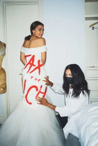 Mujer con vestido blanco de tul y la frase «Tax the rich» en rojo, mientras otra mujer le ajusta la falda, en la Met Gala